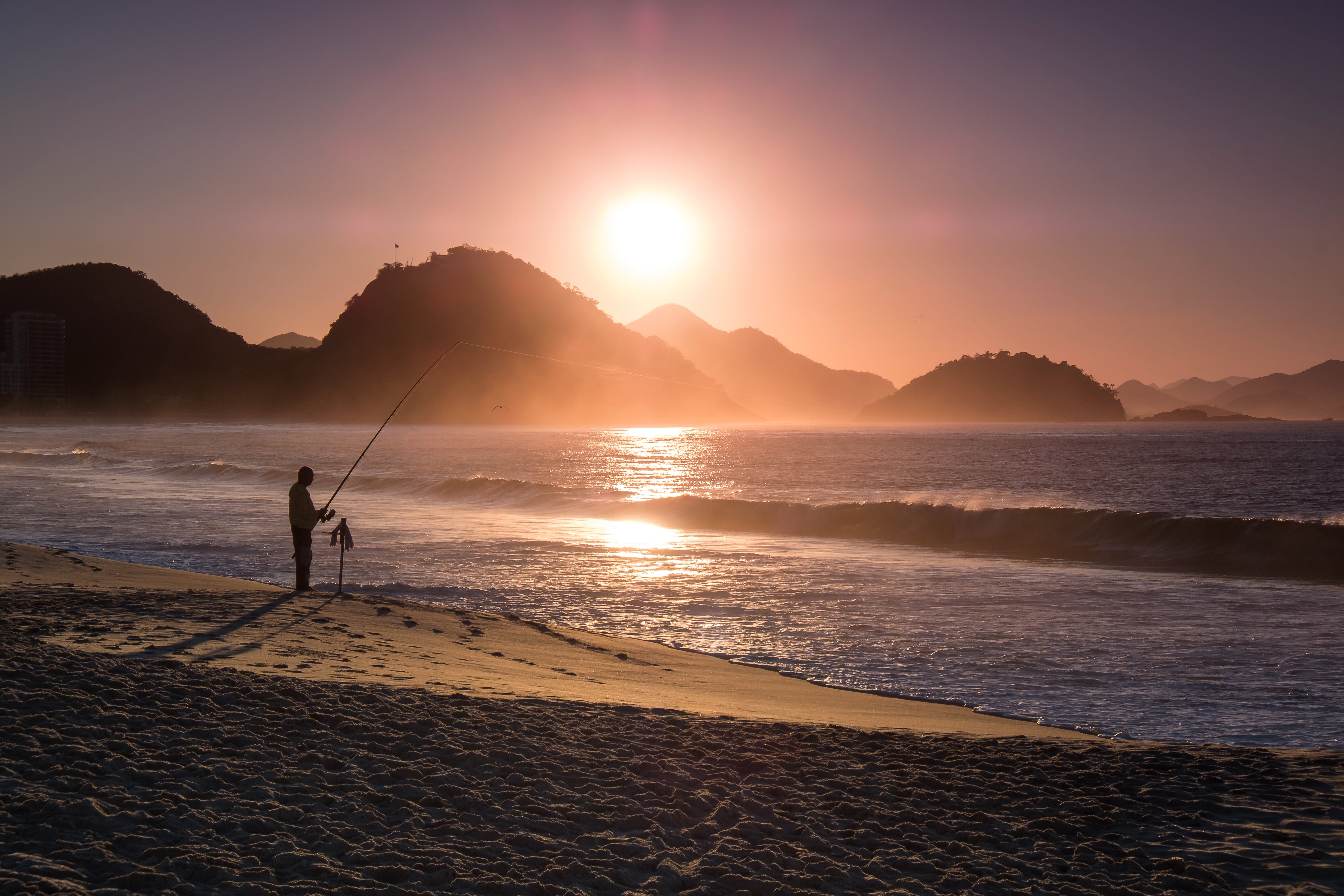 Person fishing on the beach is silhouetted at sunset